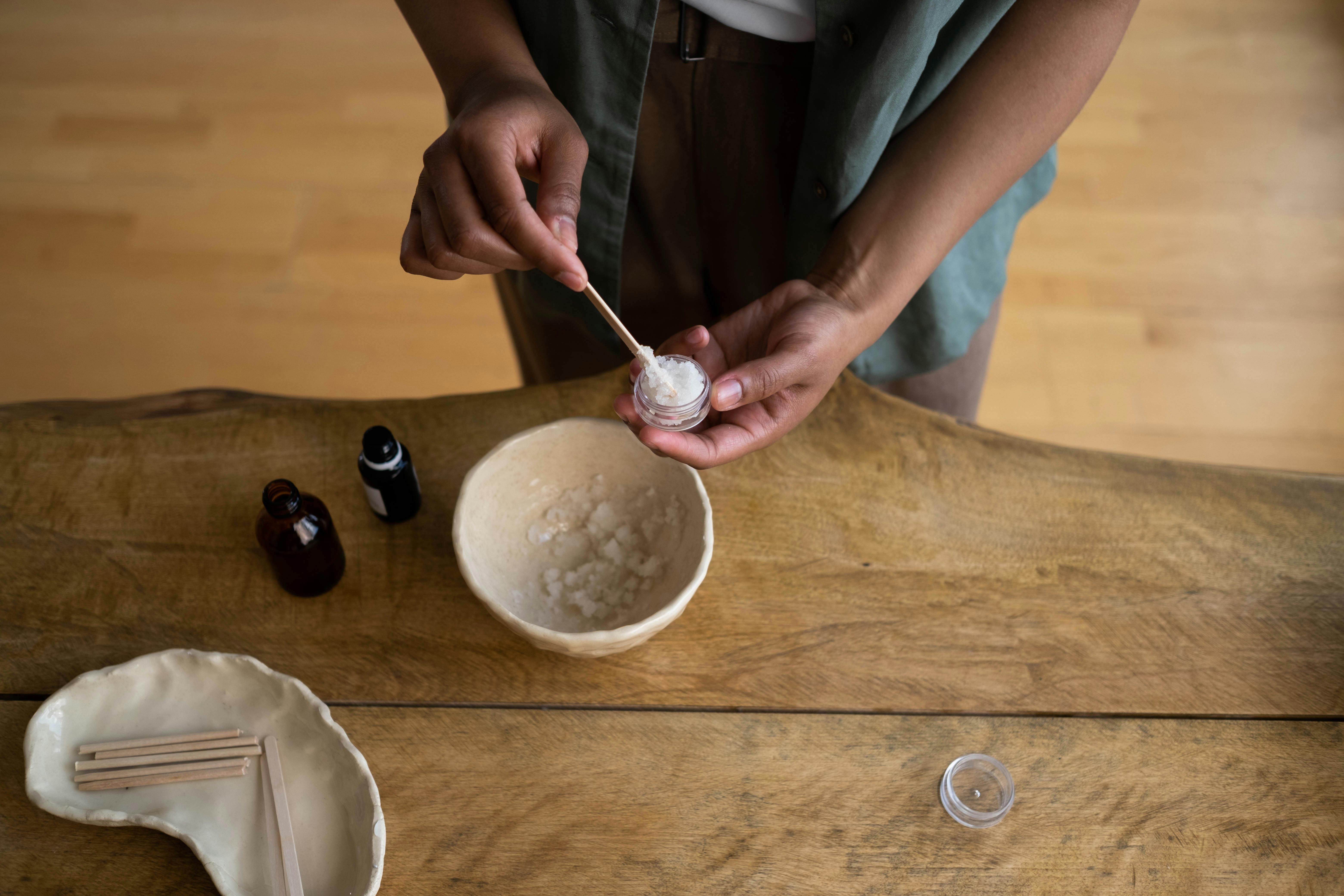 Hands transferring homemade skincare into a container on a wooden table.